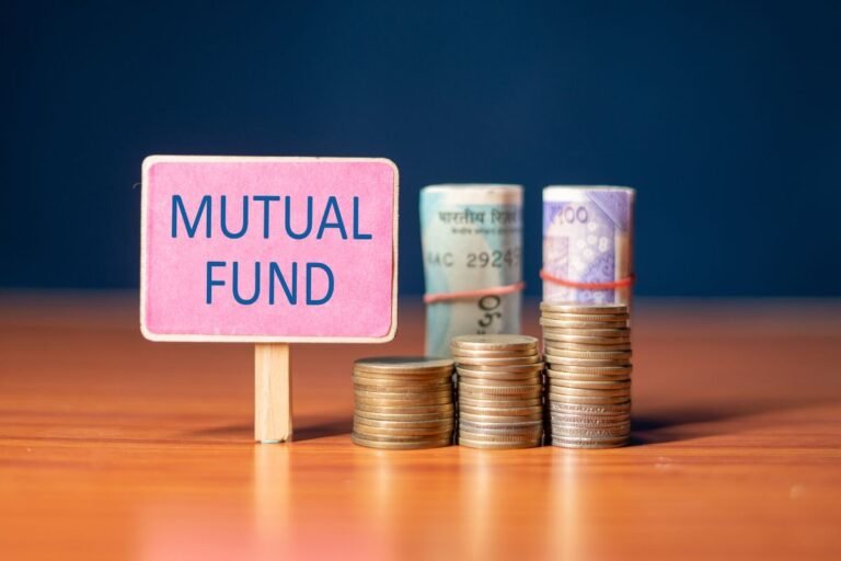 Papan tanda kecil bertuliskan MUTUAL FUND biru di depan tumpukan koin emas yang meningkat dan gulungan uang kertas Rupee India di atas meja kayu, menggambarkan pertumbuhan investasi reksa dana di pasar