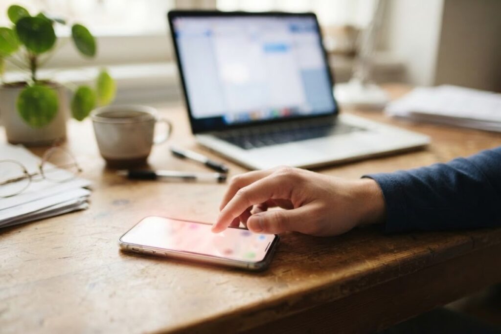 Tangan seseorang yang sedang mengetik di laptop dan memeriksa ponsel, mengilustrasikan manajemen tugas sehari-hari untuk menjaga produktivitas.